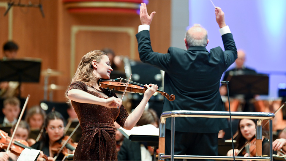 A female violinist performing in front of a conductor, conducting an orchestra behind her.